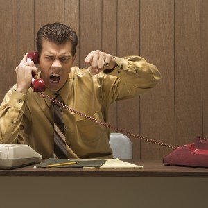Caucasion mid-adult retro businessman sitting at desk talking on telephone with angry expression and gesture.